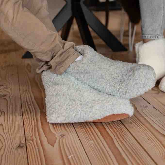 A person wearing fuzzy light gray slip-on slippers with tan soles, standing on a wooden floor. Another person's leg in beige pants is visible stepping into the slippers, and a white slipper appears on the right.