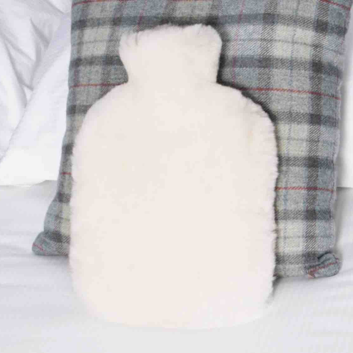 A fluffy white hot water bottle pillow placed in front of a gray and white plaid throw pillow on a white bed.