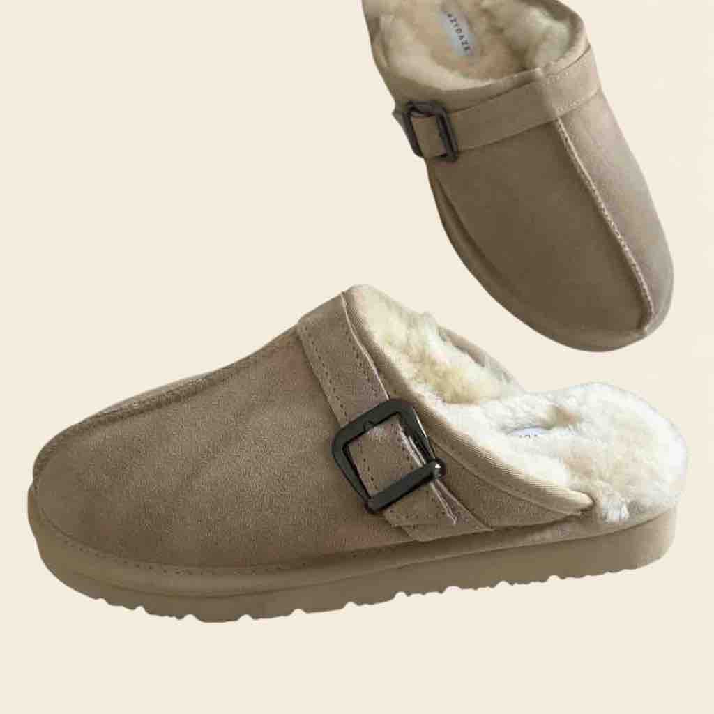 A pair of taupe suede slip-on clogs with white sherpa lining and black buckle straps, shown on a solid light beige background.