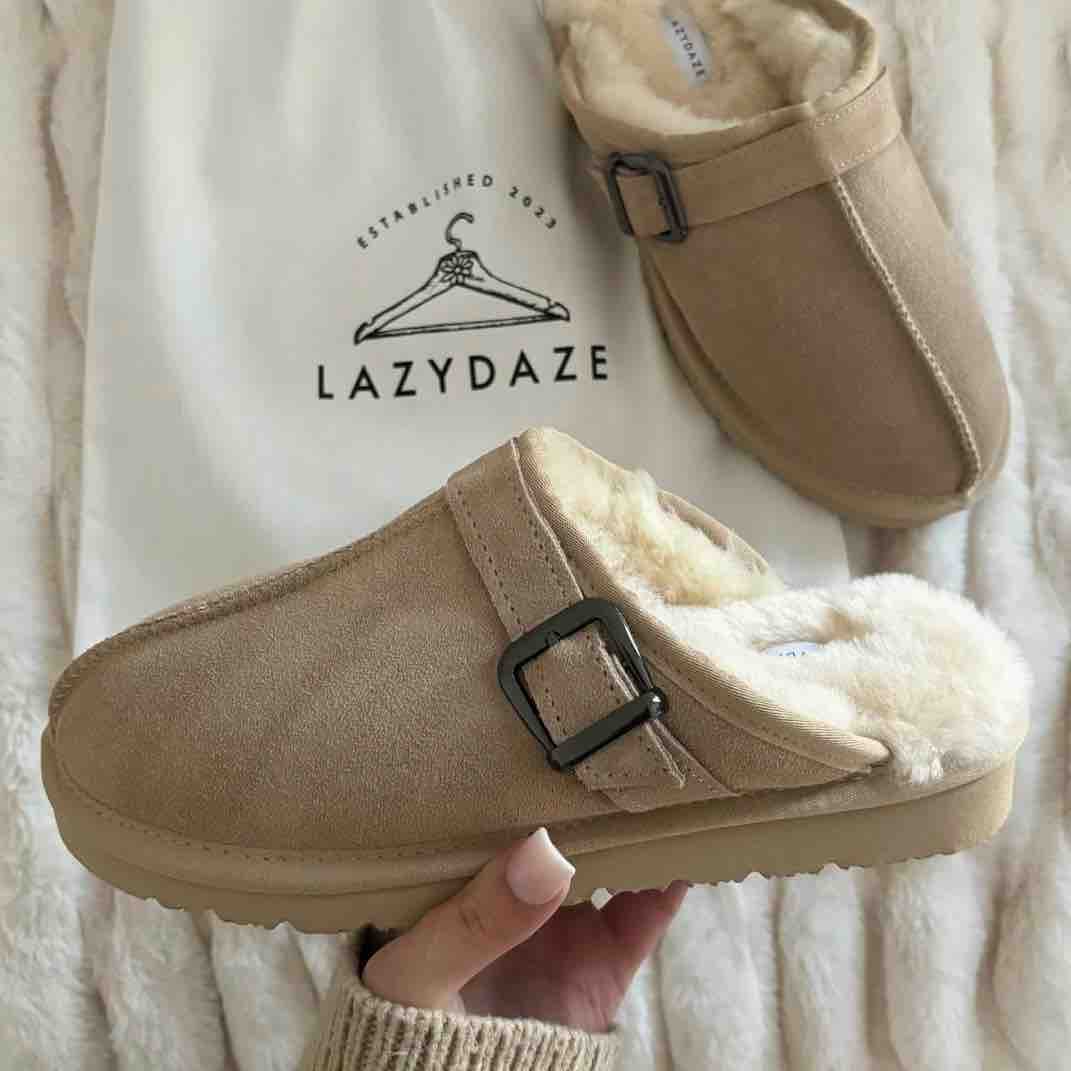 A pair of beige suede clogs with fluffy cream shearling lining and black buckle straps, shown with one shoe being held by a hand over a white textured blanket and a branded LazyDaze bag.