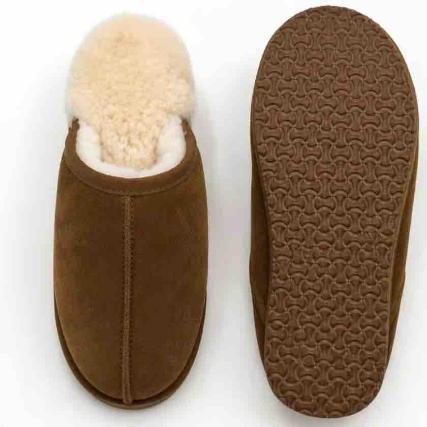 A pair of brown suede slip-on slippers shown on a white background, with one slipper viewed from the top showing a cream-colored shearling lining, and the other shown from the bottom displaying a textured brown sole.