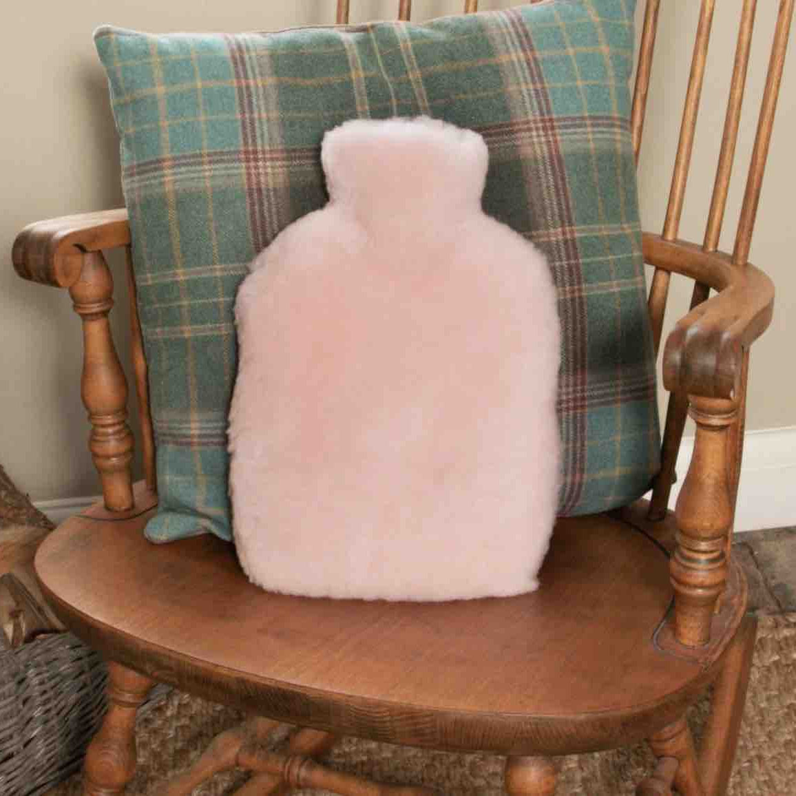 A fluffy pale pink hot water bottle cushion sits on a wooden chair in front of a green plaid throw pillow.