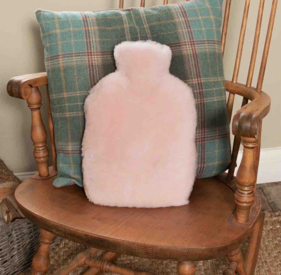 A fluffy pale pink hot water bottle cushion sits on a wooden chair in front of a green plaid throw pillow.