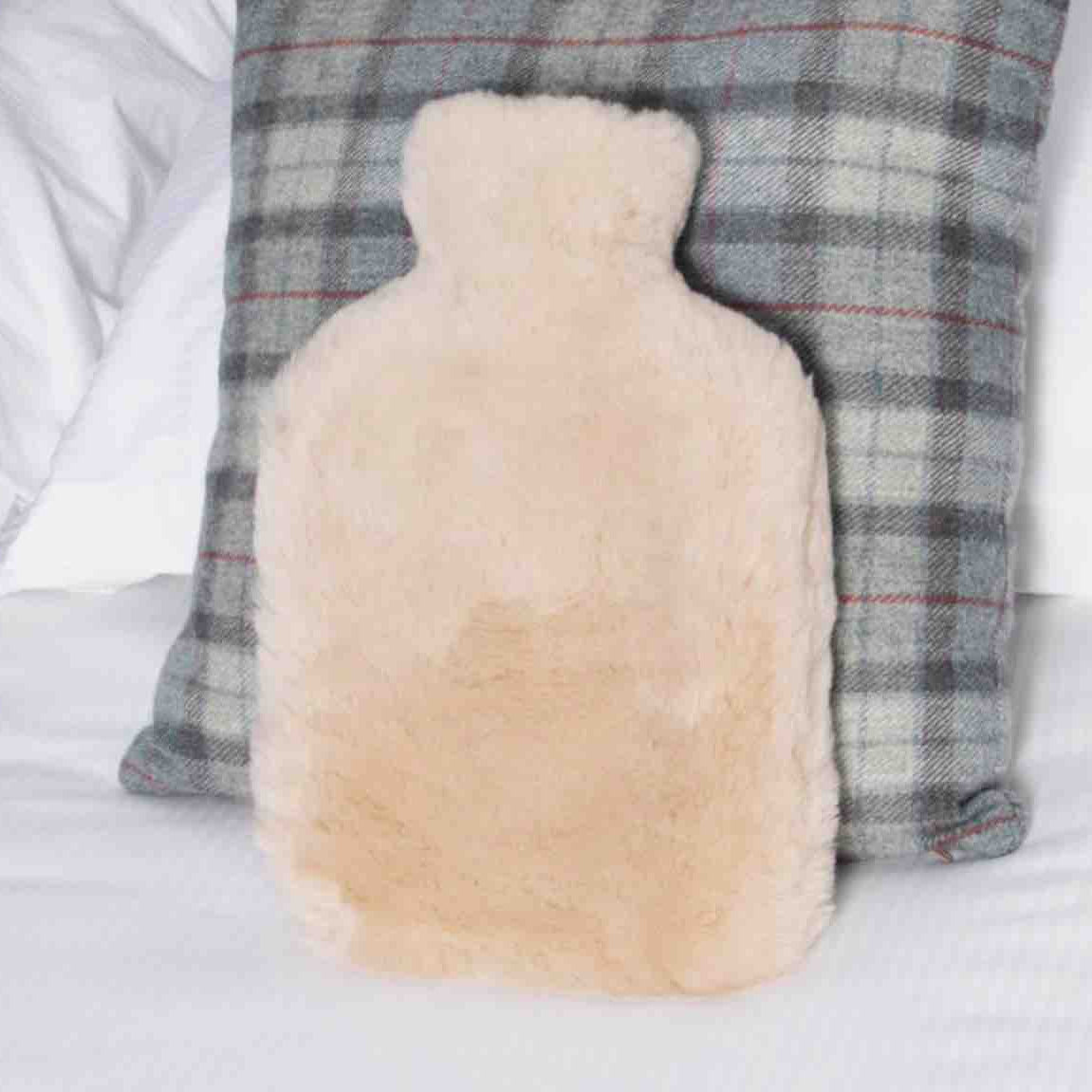 A fluffy pale beige hot water bottle pillow resting against a gray plaid pillow on a white bed.