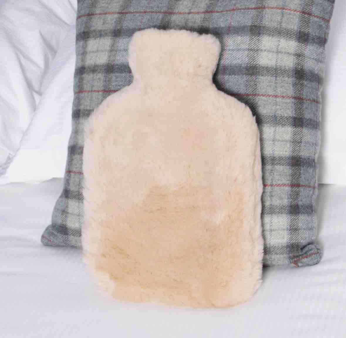 A fluffy pale beige hot water bottle pillow resting against a gray plaid pillow on a white bed.