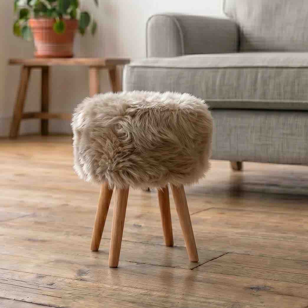 A small stool with a fluffy genuine sheepskin upholstered seat and four light wood legs, placed on a wooden floor in front of a gray sofa.
