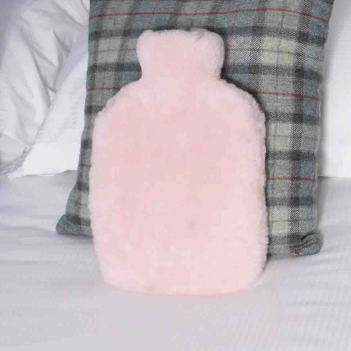 A fluffy light pink hot water bottle pillow standing upright on a white bed, positioned in front of a gray plaid pillow.