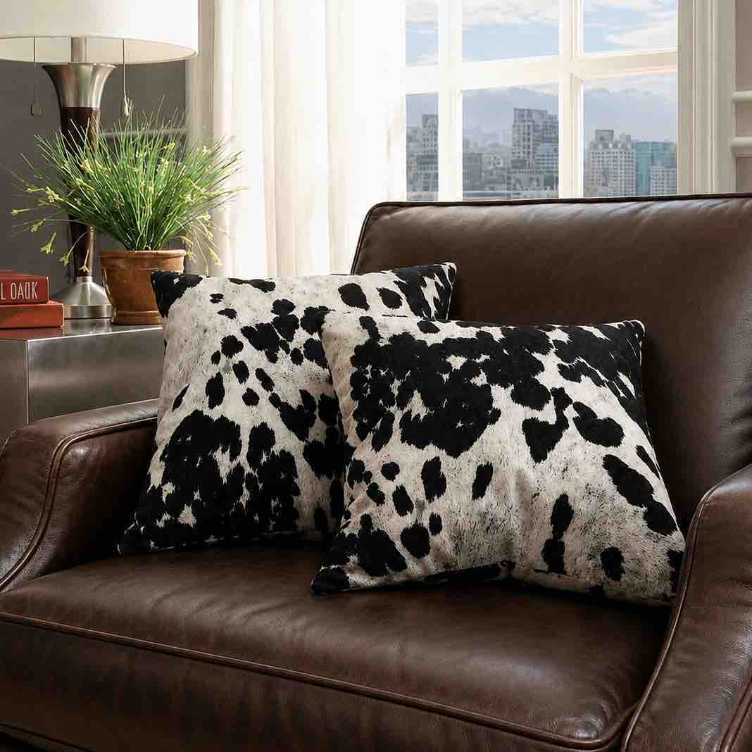 Two square throw pillows with a black and white cow print pattern resting on a brown leather sofa in a living room setting.