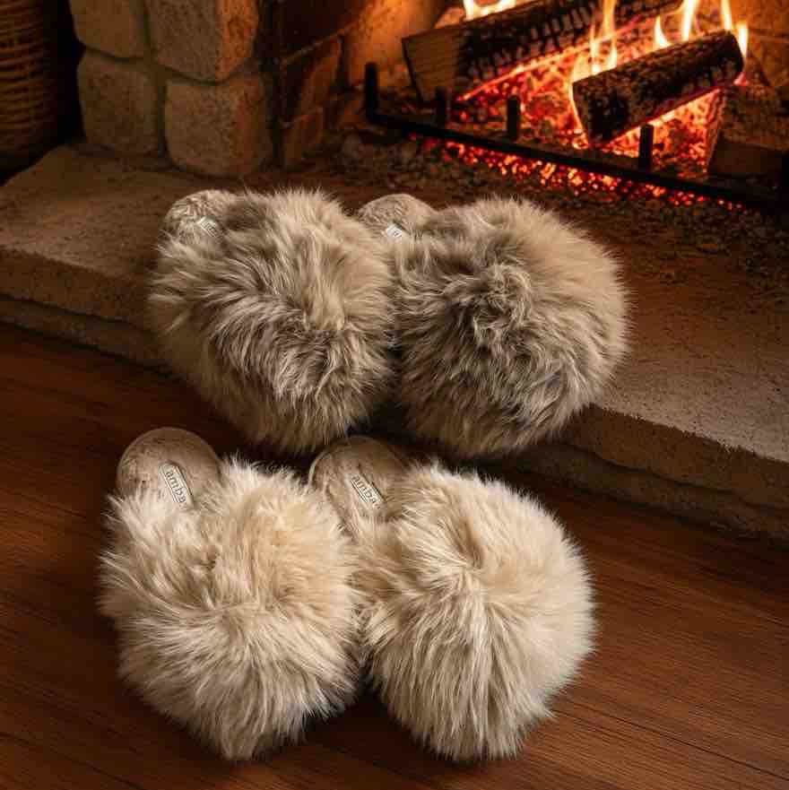 Four fluffy faux-fur slippers in light beige and cream tones arranged on a wooden floor in front of a warm fireplace with visible flames and logs.