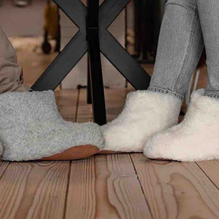A close-up of two fluffy slipper boots worn on feet, one in light gray and one in off-white, standing on a wooden floor near a black metal table leg.