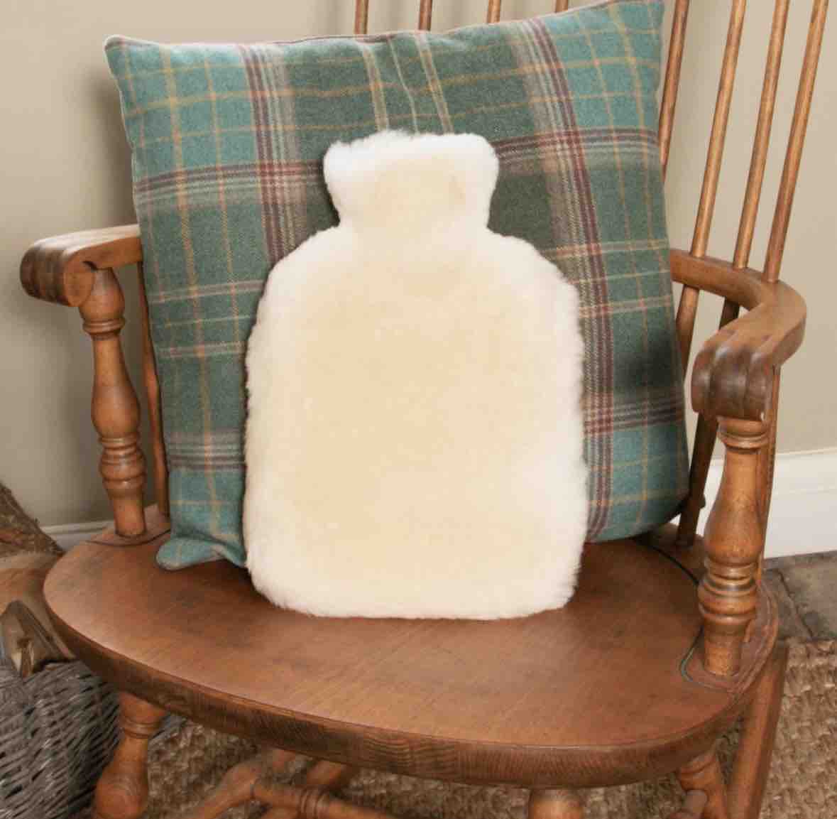 A fluffy cream-colored hot water bottle shaped like a human body, placed in front of a green plaid throw pillow on a wooden chair.