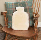 A fluffy cream-colored hot water bottle shaped like a human body, placed in front of a green plaid throw pillow on a wooden chair.
