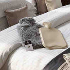 A gray fluffy hot water bottle and a beige flat hot water bottle with a neck loop, both with tags, displayed on a white striped bed alongside neutral pillows.