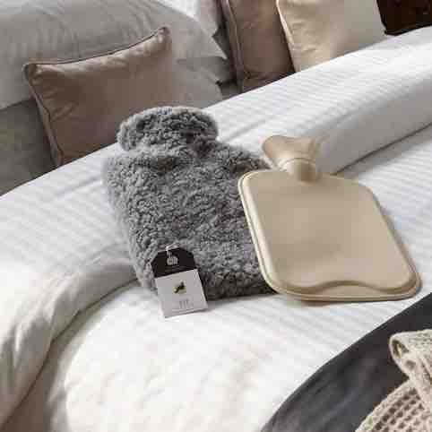 A gray fluffy hot water bottle and a beige flat hot water bottle with a neck loop, both with tags, displayed on a white striped bed alongside neutral pillows.