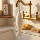 A fish-shaped soap on a rope hanging from a brass faucet in a bathroom setting, shown within a mobile browser interface. The soap is textured and cream-colored, with a warm beige tiled background and a gold-toned faucet.