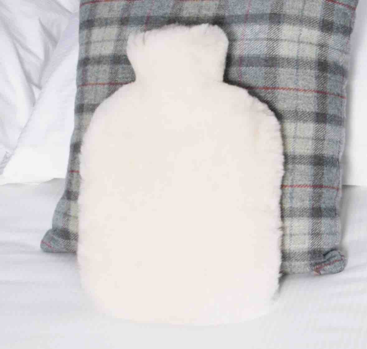 A fluffy white hot water bottle pillow placed in front of a gray and white plaid throw pillow on a white bed.