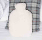 A fluffy white hot water bottle pillow placed in front of a gray and white plaid throw pillow on a white bed.