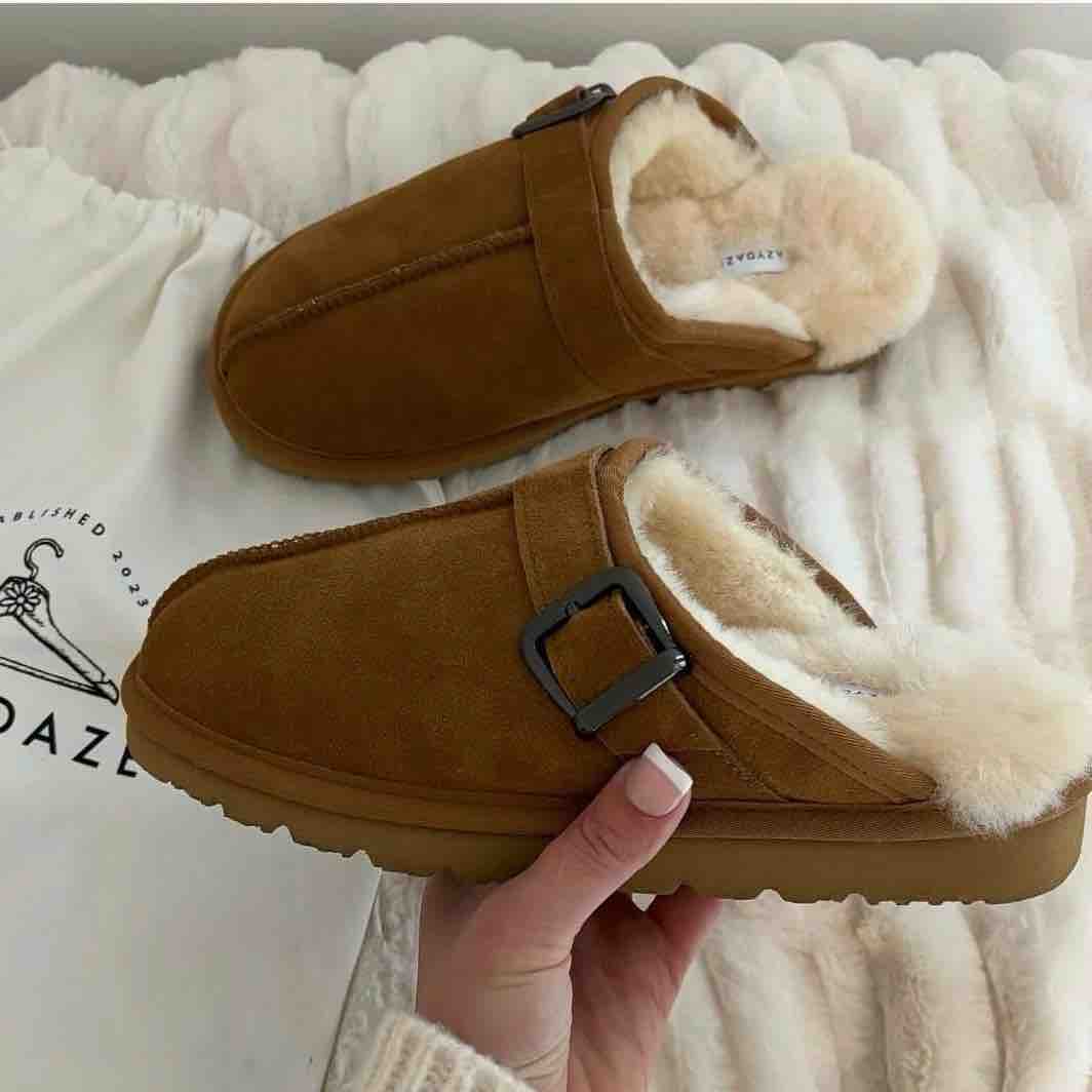 A pair of brown suede clog-style slippers with cream faux-fur lining and a dark buckle strap, shown with one being held by a hand over a white textured blanket and branded drawstring bag.