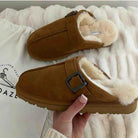A pair of brown suede clog-style slippers with cream faux-fur lining and a dark buckle strap, shown with one being held by a hand over a white textured blanket and branded drawstring bag.
