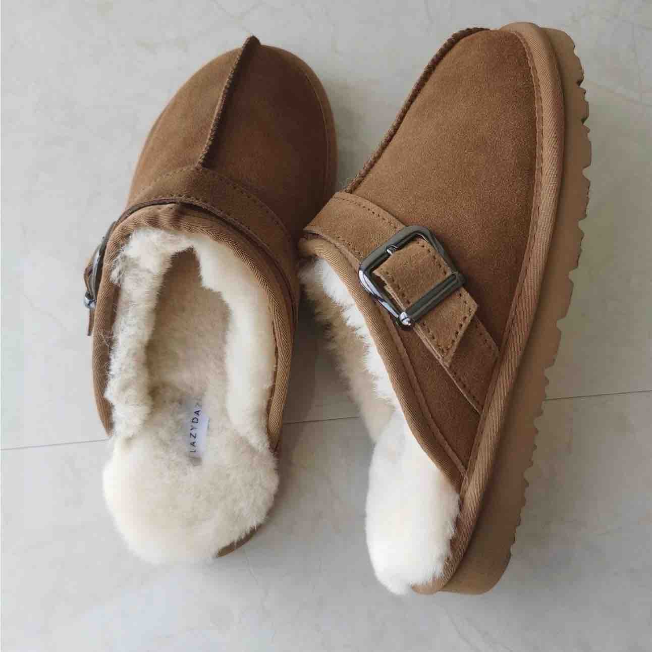 A pair of tan suede slip-on slippers with white faux-fur lining and a silver buckle detail, shown on a light gray tiled floor.