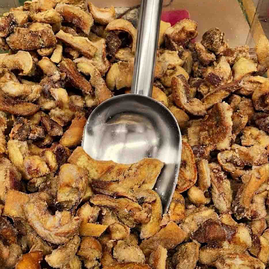 A close-up of dried, curled slices of what appears to be dried banana or plantain, piled in a clear container with a large metal scoop resting in the center.