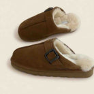 A pair of brown suede slip-on clogs with a soft white shearling lining and a black buckle detail, displayed on a light beige background.