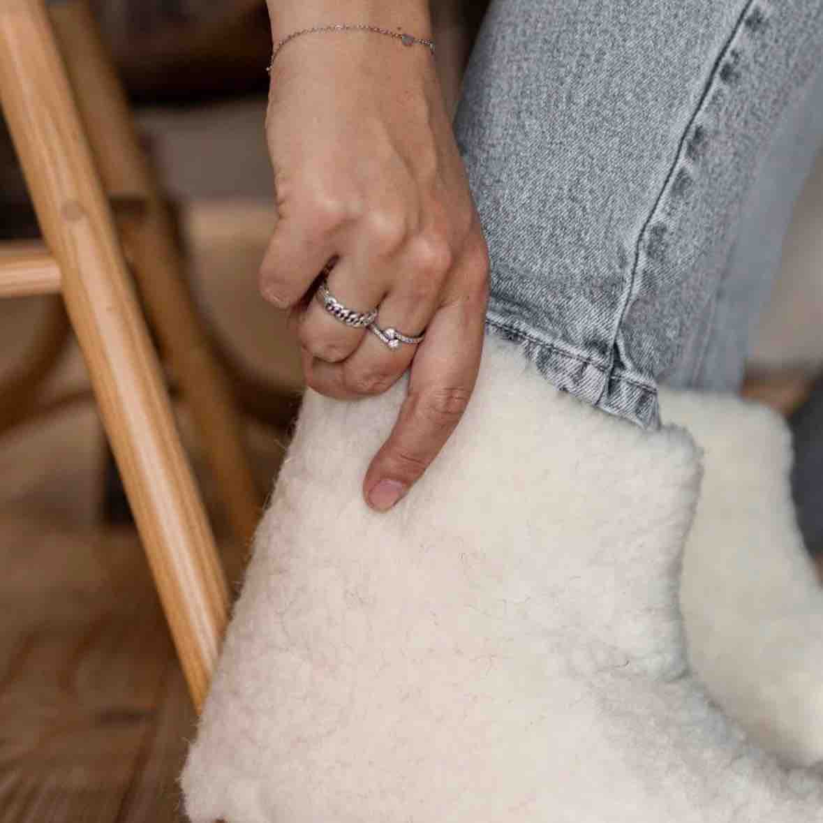 A close-up lifestyle shot of a person's hand resting on a fluffy off-white sheepskin or faux-shearling boot, wearing silver rings and a delicate bracelet, paired with light-wash denim jeans.