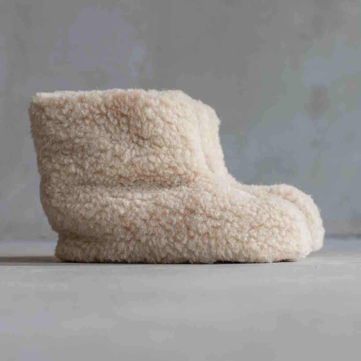 A single plush, ankle-height slipper in a warm cream or off-white sherpa/fleece material, photographed against a soft gray studio background.