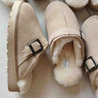 A pair of beige suede slip-on slippers with fluffy cream shearling lining and black buckle straps, shown on a light neutral background.
