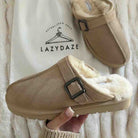 A pair of beige suede clogs with fluffy cream shearling lining and black buckle straps, shown with one shoe being held by a hand over a white textured blanket and a branded LazyDaze bag.