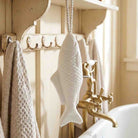 A white fish-shaped soap hanging from a rope hook on a rustic wooden shelf in a bathroom setting with a brass faucet and textured towels.