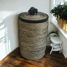 Woven storage basket with a black lid on a wooden floor, with a plant in the background.