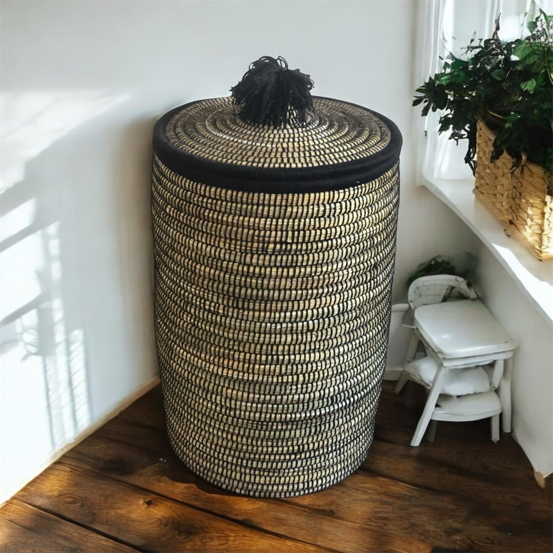 Woven storage basket with a black lid on a wooden floor, with a plant in the background.