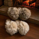 Four fluffy faux-fur slippers in light beige and cream tones arranged on a wooden floor in front of a warm brick fireplace with glowing embers.