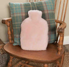 A fluffy pale pink hot water bottle cushion sits on a wooden chair in front of a green plaid throw pillow.