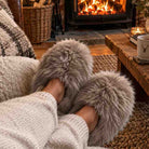 A cozy close-up of a person wearing fluffy gray faux-fur slippers while sitting on a couch in a warm living room with a lit fireplace and wood coffee table in the background.