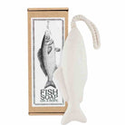 A fish-shaped white soap bar with a rope loop, shown next to its brown cardboard packaging box featuring a black-and-white fish illustration and the text 'FISH SOAP ON A ROPE'.