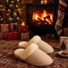 A pair of cream-colored fleece slip-on slippers with white sherpa lining, placed on a patterned rug in front of a cozy fireplace with a burning wood stove, a lit Christmas tree, wrapped gifts, and a Christmas stocking in the background.