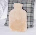 A fluffy pale beige hot water bottle pillow resting against a gray plaid pillow on a white bed.