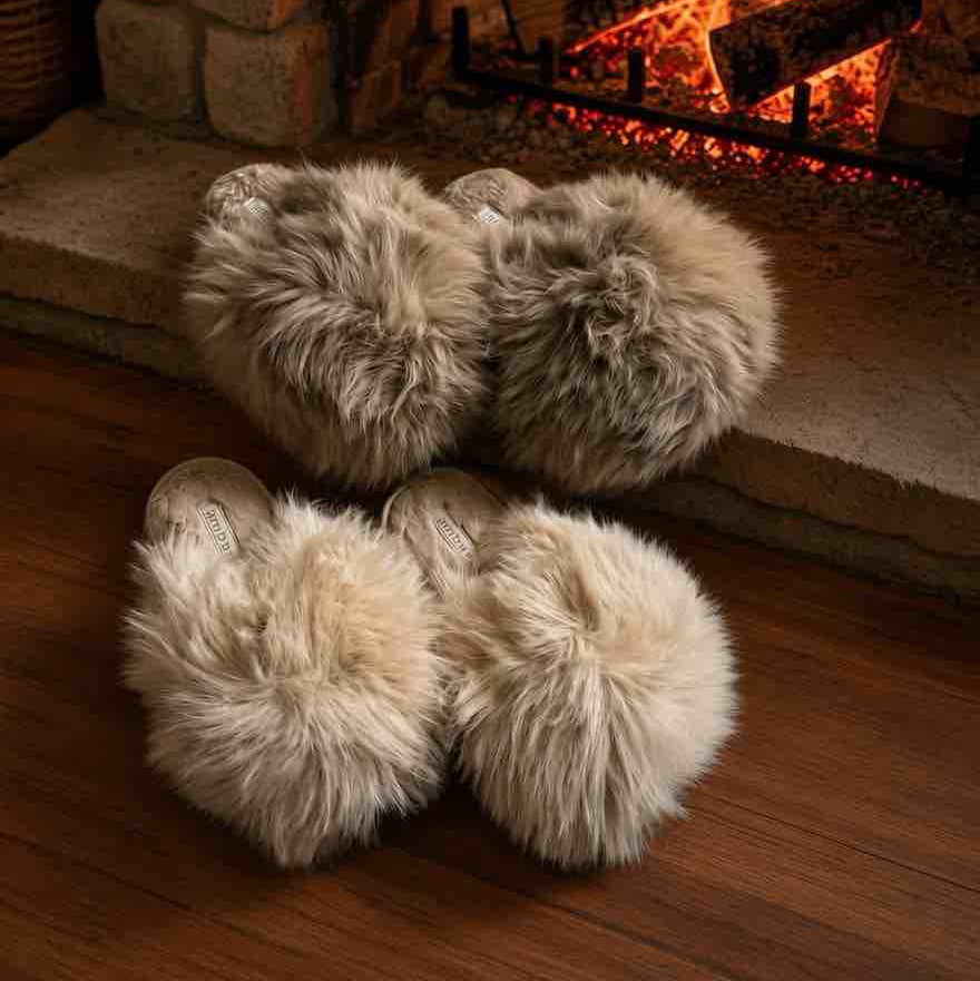 Four fluffy faux-fur slippers in light beige and cream tones arranged on a wooden floor in front of a warm brick fireplace with glowing embers.