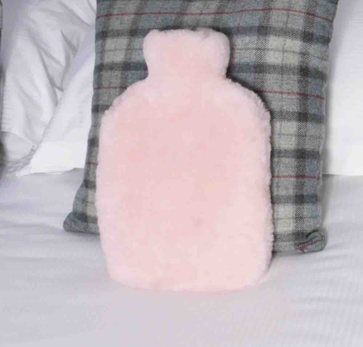 A fluffy light pink hot water bottle pillow standing upright on a white bed, positioned in front of a gray plaid pillow.