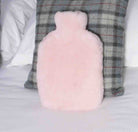 A fluffy light pink hot water bottle pillow standing upright on a white bed, positioned in front of a gray plaid pillow.