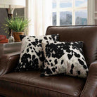Two square throw pillows with a black and white cow print pattern resting on a brown leather sofa in a living room setting.