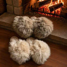 Four fluffy faux-fur slippers in light beige and cream tones arranged on a wooden floor in front of a warm fireplace with visible flames and logs.